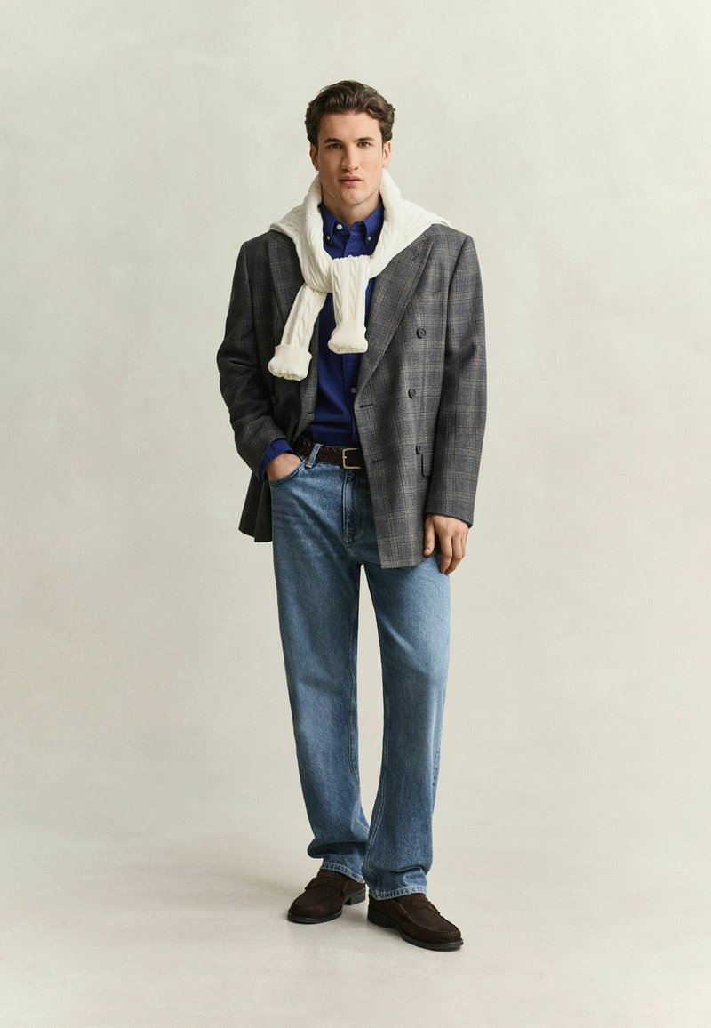 Junger Mann steht mit einer Hand in der Tasche, trägt einen grauen karierte Blazer, ein blaues Hemd, hellblaue Jeans, braune Schuhe und einen weißen Pullover um die Schultern.