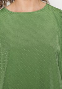 Top verde de manga curta com superfície texturizada de pequenos pontos salientes, apresentando um decote arredondado e um caimento suave.
