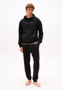 Čierna mikina s kapucňou a predným vreckom, bielym logom, doplnená o čierne nohavice jogger s bočným vreckom a logom na nohe.