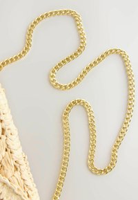 Chaîne épaisse de ton or attachée à un sac beige tissé, présenté sur une surface gris clair.