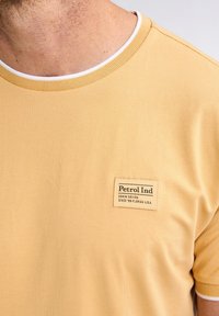 Tricou galben cu mânecă scurtă, fabricat din bumbac, cu guler rotund, având un eticheta cusută pe piept cu numele brandului și detalii.