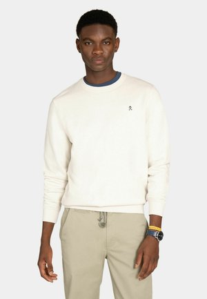 Joven con sudadera blanca, pantalones beige, reloj azul y pulsera amarilla, de pie frente a un fondo claro y sencillo.