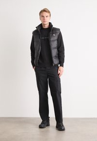 Gilet imbottito nero sopra una felpa nera, abbinato a pantaloni cargo neri e scarpe nere. Dotato di chiusura a zip e dettaglio con logo.