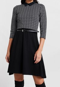 Robe noire avec un haut ajusté arborant un motif en losange blanc, un col, des manches trois-quarts et une ceinture noire à la taille.