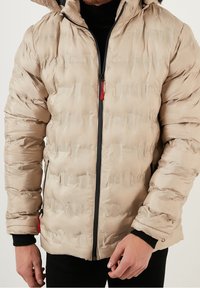 Beige puffer jacket with a textured finish, featuring a black zipper and red accents, adjustable hood, and elastic cuffs.