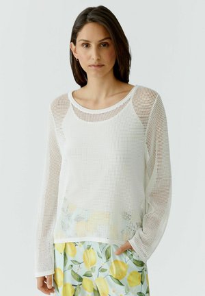 Vrouw draagt een wit mesh top met lange mouwen over een wit hemdje, gecombineerd met een broek met een geel citroen- en groen bladmuster.