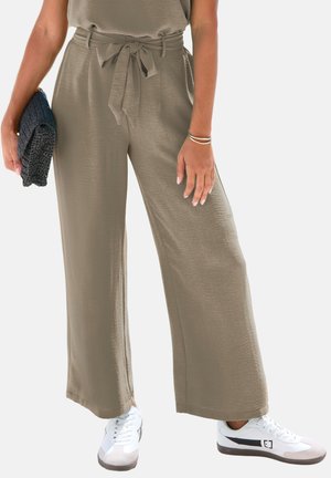 Taupe weit geschnittene Hose mit selbstgebundenem Gürtel, kombiniert mit weißen Turnschuhen, goldenen Armbändern und einer schwarzen gewebten Clutch, die in der linken Hand gehalten wird.