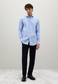 Chemise bleu clair à manches longues en tissu lisse, assortie à un pantalon marine foncé et des chaussures noires. La chemise possède un col standard.