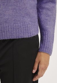 Lilla sweater med en blød, fuzzy tekstur. Ribbet manchetter og kant. Isoleret sammen med sorte bukser, viser et nærbillede af ærmet og hånden.