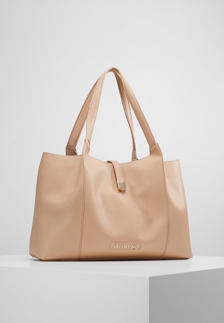 Beige Valentino läder tote-väska med dubbla handtag, en framslutning med rem och en guldlogoplatta på en vit pedistal.