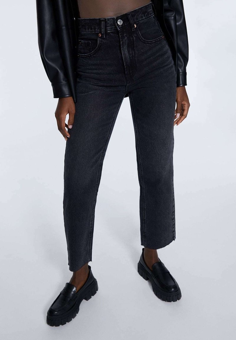 Jeans en denim noir taille haute avec une coupe évasée, présentant des poches avant et arrière, ainsi que des détails de couture subtils. Portés avec des mocassins noirs.