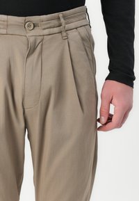 Beige Hosen mit Falten, mit Knopfverschluss und Seitentaschen. Aus leichtem Stoff mit glatter Textur gefertigt.
