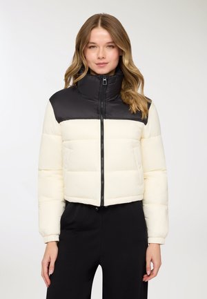 Piumino corto con sezione superiore nera e sezione inferiore crema, caratterizzato da un colletto alto e chiusura a zip. Texture morbida con design trapuntato.