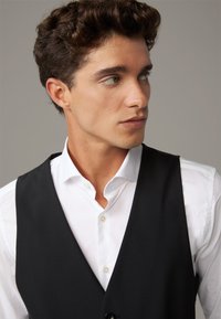 Gilet nero sopra una camicia bianca con bottoni. Il gilet ha una superficie liscia e un taglio classico, con una chiusura a un bottone e senza motivi visibili.