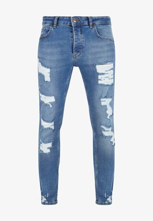 Karcsú kék farmernadrág, kopott részletekkel, több hasítékkal és szétfoszlott szélekkel, öt zsebbel és elülső gombos zárással.