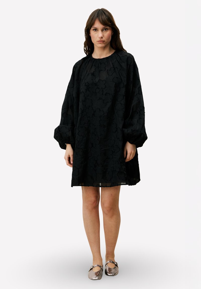 Robe noire oversize avec manches bouffantes, motif floral texturé et col rond. Associée à des chaussures plates brillantes.