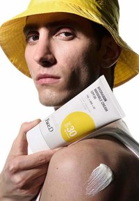 Tubetto di crema solare etichettato "Hydrasum Invisible Cream SPF 30" tenuto da una figura. Cappuccio giallo e crema bianca spalmata su una spalla e un braccio nudi.