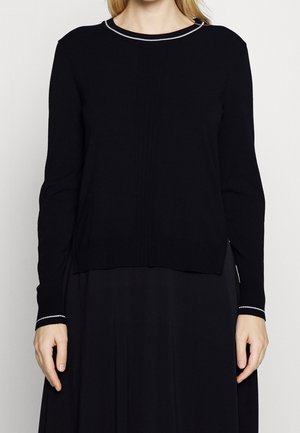 Woman wearing a black long-sleeve sweater with white trim on collar and cuffs, paired with a black skirt.