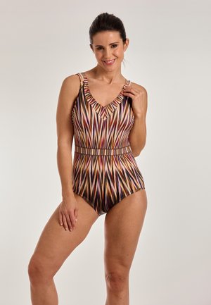 Naeratav naine, kandes ühes tükis ujumistrikood mitmevärviliste vertikaalsete triipudega ja voldilise kaelusega, seisab ühevärvilise heleda tausta ees.