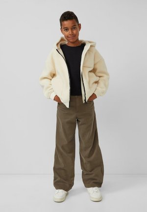 Crème faux fur hooded jack met een ritssluiting aan de voorkant, een zwart shirt eronder, gecombineerd met olijfgroene broek en witte sneakers.