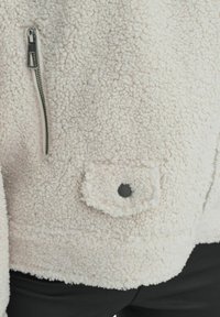 Giacca in pile bianca con superficie strutturata e pelosa, zip argentata e una piccola tasca frontale con dettaglio del bottone nero.