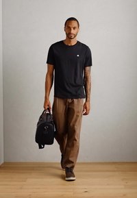 T-shirt en coton noir, pantalon marron ample et baskets grises. Porte un sac en cuir noir avec des accents de quincaillerie contrastants.
