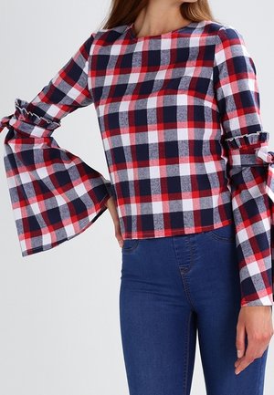 Kvinde iført rød, hvid og marineblå ternet langærmet top med flagrende ærmer og bundet detaljer, kombineret med højtaljede blå jeans.