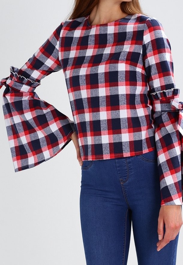 Femme portant un haut à manches longues à carreaux rouge, blanc et bleu marine avec des manches évasées et des détails noués, associé à un jean bleu taille haute.