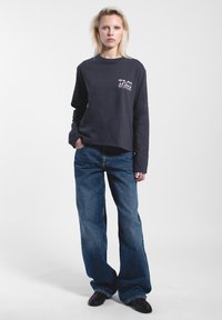 Schwarzes Langarmshirt mit einem kleinen Grafiklogo auf der Brust, kombiniert mit weit geschnittenen blauen Jeans und schwarzen Schuhen. Einfarbiger Hintergrund.