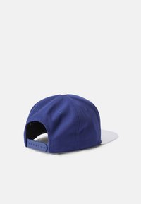 Gorra de béisbol de tela azul con visera plana blanca. Presenta un cierre ajustable tipo snapback, detalles de costura y agujeros de ventilación a los lados.