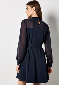 Vestido de marinero con una falda suave, mangas largas semitransparentes y un cuello con abalorios. Incluye una abertura en la parte posterior con forma de llave y un cinturón de tela.