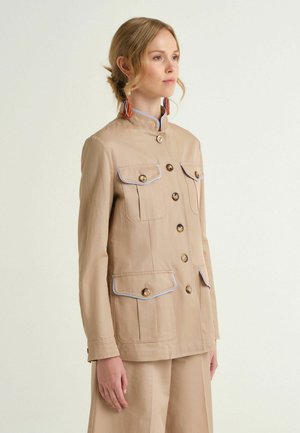 Femme portant une veste beige boutonnée avec quatre poches avant et des bordures contrastantes, associée à un pantalon large assorti.