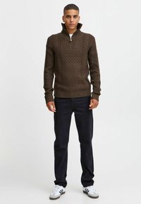 Suéter marrón de punto con cuello alto, que presenta un patrón texturizado. Combinado con jeans oscuros y zapatillas blancas.