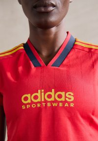 Sarkanais Adidas sporta tērps ar V veida dekoltē, kas ir apzīmēts ar dzelteniem logo un svītrām, kā arī ar teksturētu audumu ar vertikālām līnijām.