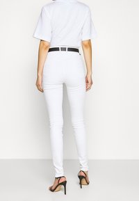 Jeans blancos ajustados con un diseño de cintura alta, bolsillos traseros y un cinturón negro. La modelo lleva sandalias negras de tacón alto.