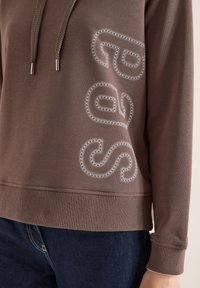 Gros plan sur un sweat à capuche marron avec cordons de serrage et lettres "SPORT" cousues sur le côté, assorti à un jean bleu foncé.