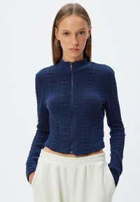 Mörkblå stickad zip-up tröja med hög krage, med ett strukturerat mönster, smal passform och långa ärmar. Bärs med ljusa byxor.