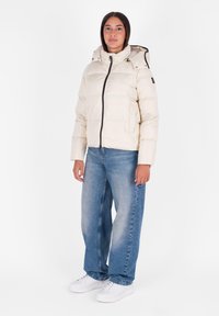 Kremowa kurtka puchowa z kapturem, z zamkiem błyskawicznym z przodu i widocznym przeszyciem. Połączona z jasnoniebieskimi spodniami typu wide-leg i białymi trampkami.