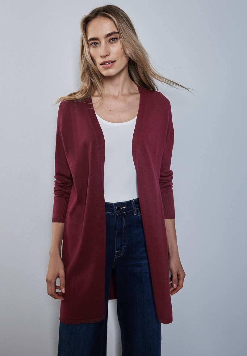Burgundygrauer langer Cardigan aus weichem Stoff, mit V-Ausschnitt und offener Vorderseite. Kombiniert mit einem weißen Oberteil und blauen Jeans.