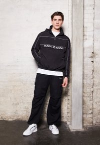 Sort zip-up jakke med hvid syning og logo, parret med sorte cargo bukser. Modellen bærer hvide sneakers, sat mod en neutral baggrund.