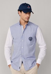 Uomo con camicia a righe blu e bianche, maniche bianche, pantaloni beige e cappellino da baseball blu navy che guarda leggermente verso destra.