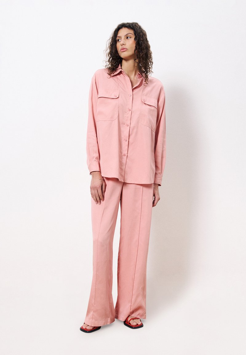 Pink, lightweight, long-sleeve shirt with two chest pockets and button closure, paired with matching wide-leg pants and black sandals.