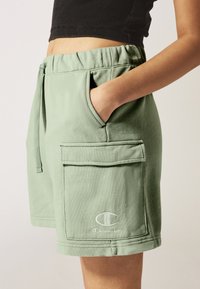 Pantaloni corti verde chiaro con vita elastica, tasche laterali e tasca frontale a toppa. Presenta il logo Champion ricamato in bianco.