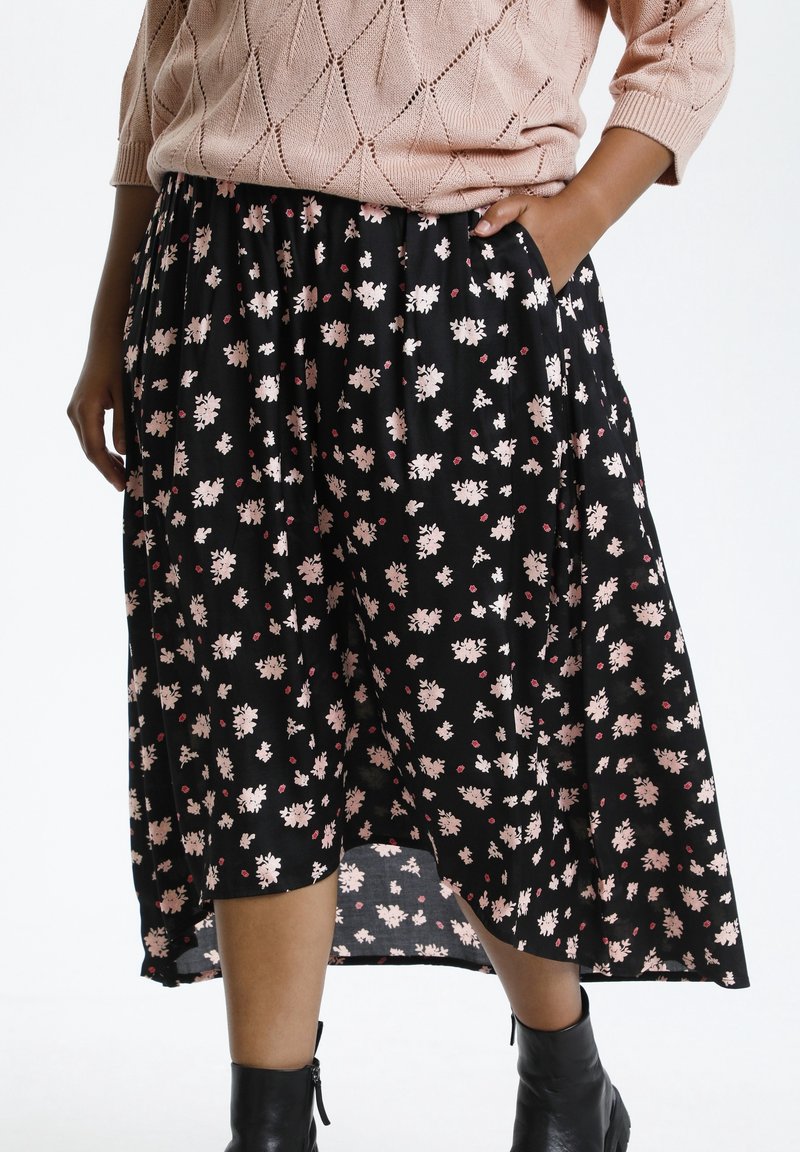 Femme portant une jupe midi noire à fleurs avec des poches, un pull en maille rose et des bottines noires, debout avec une main dans la poche.