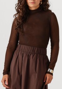 Bruine langslieve top van transparant materiaal met een hoge hals, gecombineerd met een donkerbruine rok met een gathered taille. Bevat een zilveren armband.
