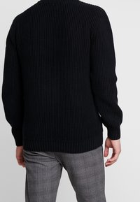 Czarny dziergany sweter z teksturowanym, prążkowanym wzorem. Posiada długie rękawy i dopasowany pas. Połączony z szarymi spodniami w kratę.