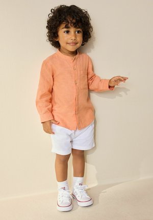 Malé dítě s kudrnatými vlasy v broskvovém tričku s dlouhým rukávem, bílých šortkách, bílých ponožkách a bílých teniskách, stojí u béžové zdi.