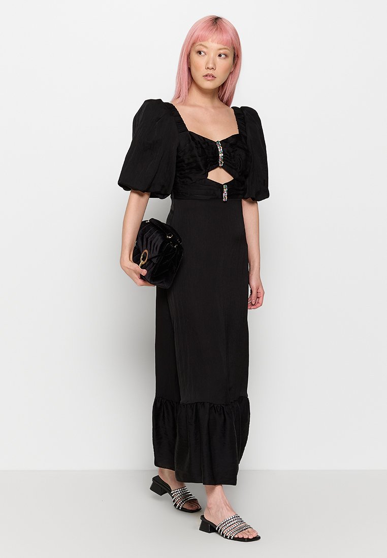 Schwarzes Kleid mit Puffärmeln, einem Ausschnitt und einem gerafften Saum. Kombiniert mit einer schwarzen runden Tasche und klobigen Sandalen mit Absätzen und Verzierungen.