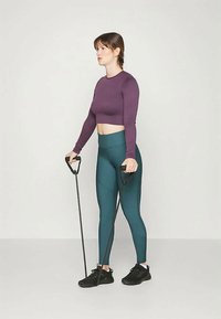 Fekete ellenállás-szalaggal edző nő lila hosszú ujjú crop toppban és türkiz leggingsben egy sima világos háttér előtt.