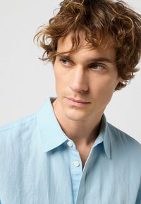 Chemise bleu clair à manches longues en tissu doux, dotée d'un col pointu, d'une texture subtile et d'une fermeture à un bouton.